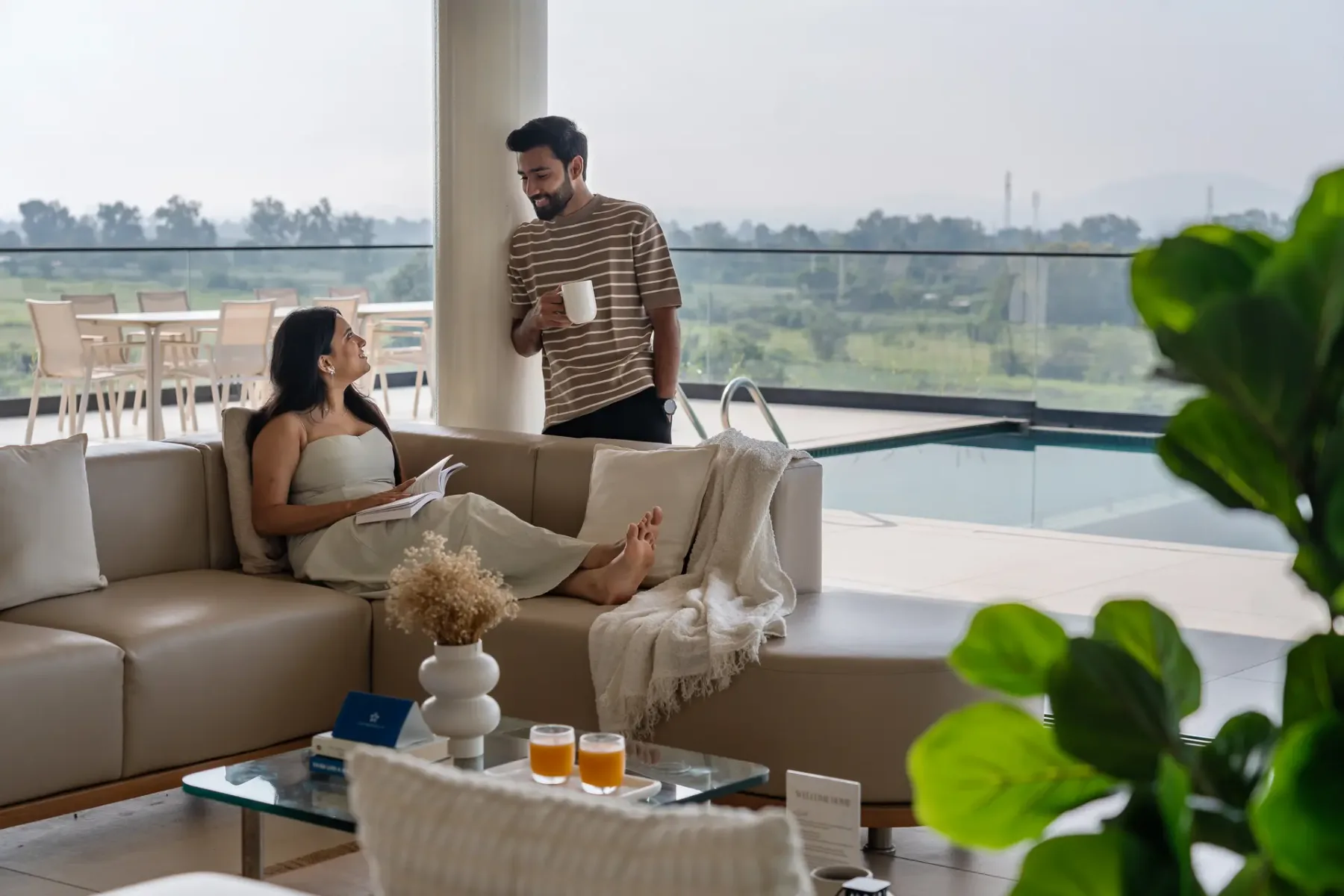 Una sala de estar con dos personas relajándose junto a grandes ventanales y una piscina.