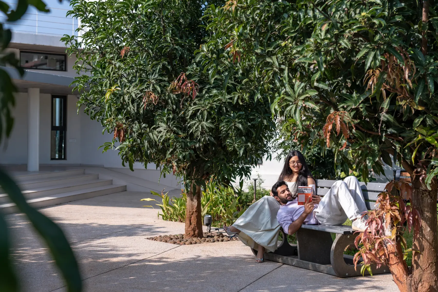 Una pareja descansa en un asiento exterior bajo árboles frondosos junto a un edificio moderno.