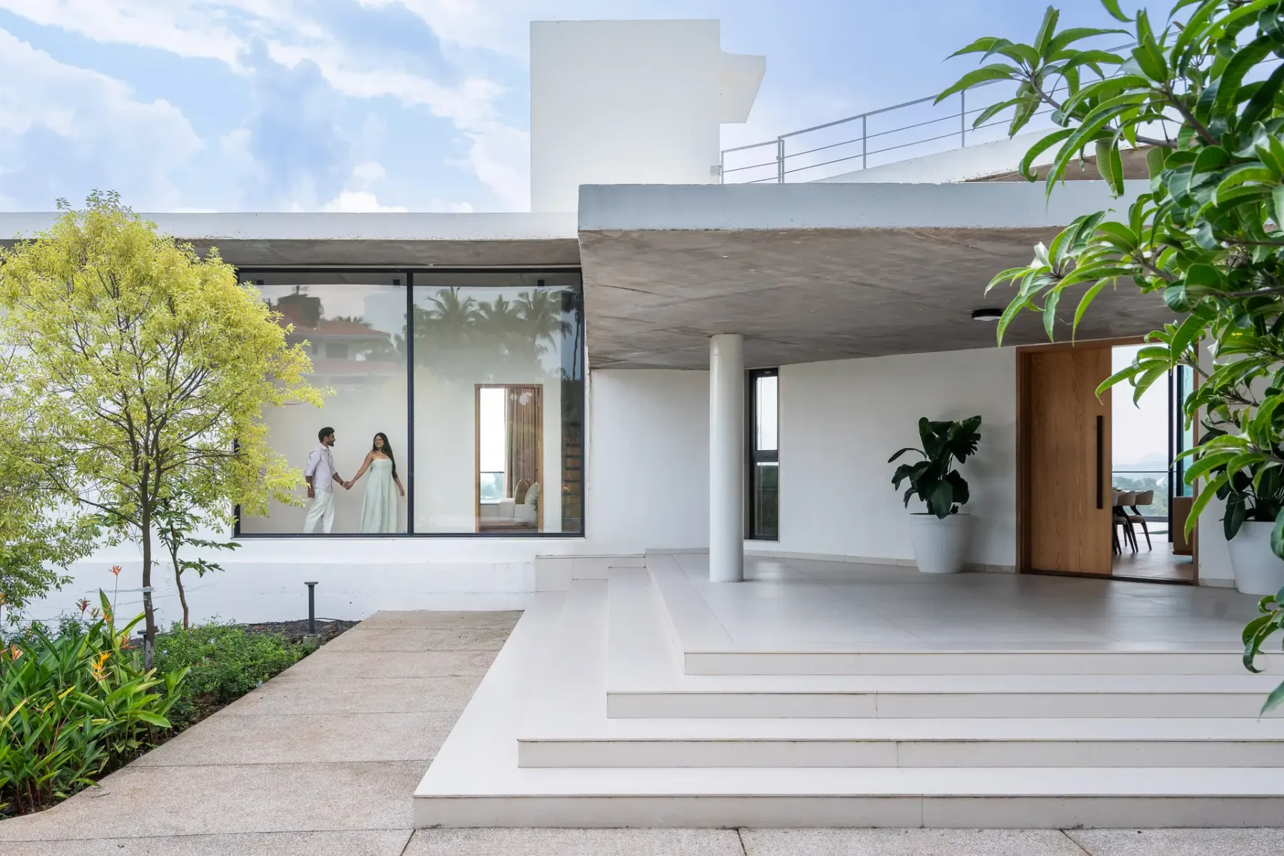 Entrada de una casa moderna con amplios escalones blancos, ventanas de vidrio y plantas alrededor.