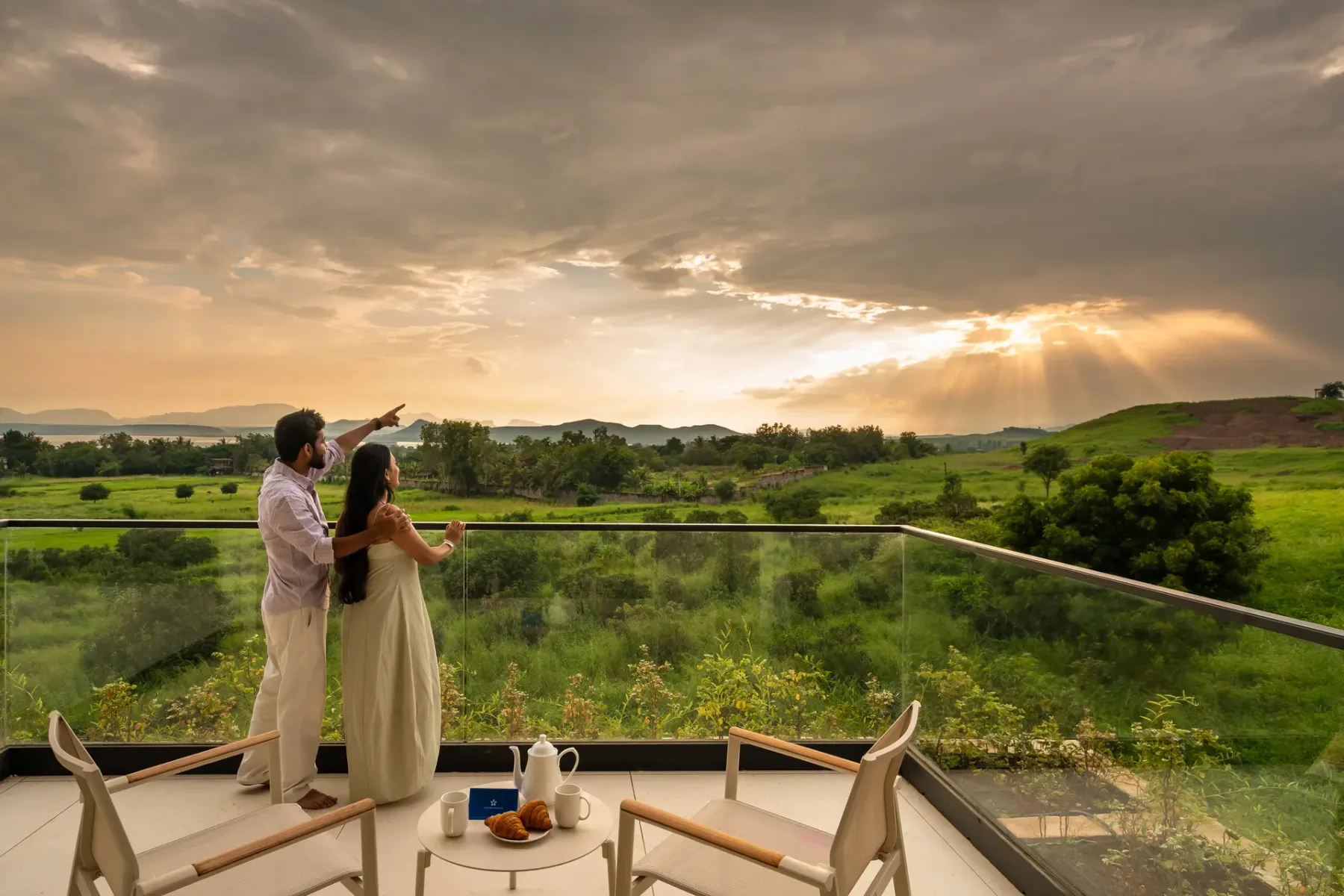 Una pareja está junto a una barandilla de vidrio con vistas a colinas verdes al atardecer.