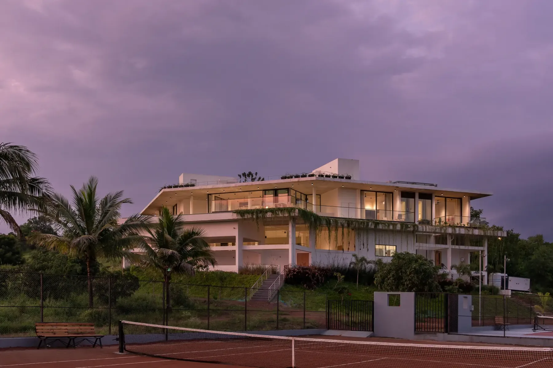 Un edificio moderno da a una cancha de tenis al aire libre al anochecer.