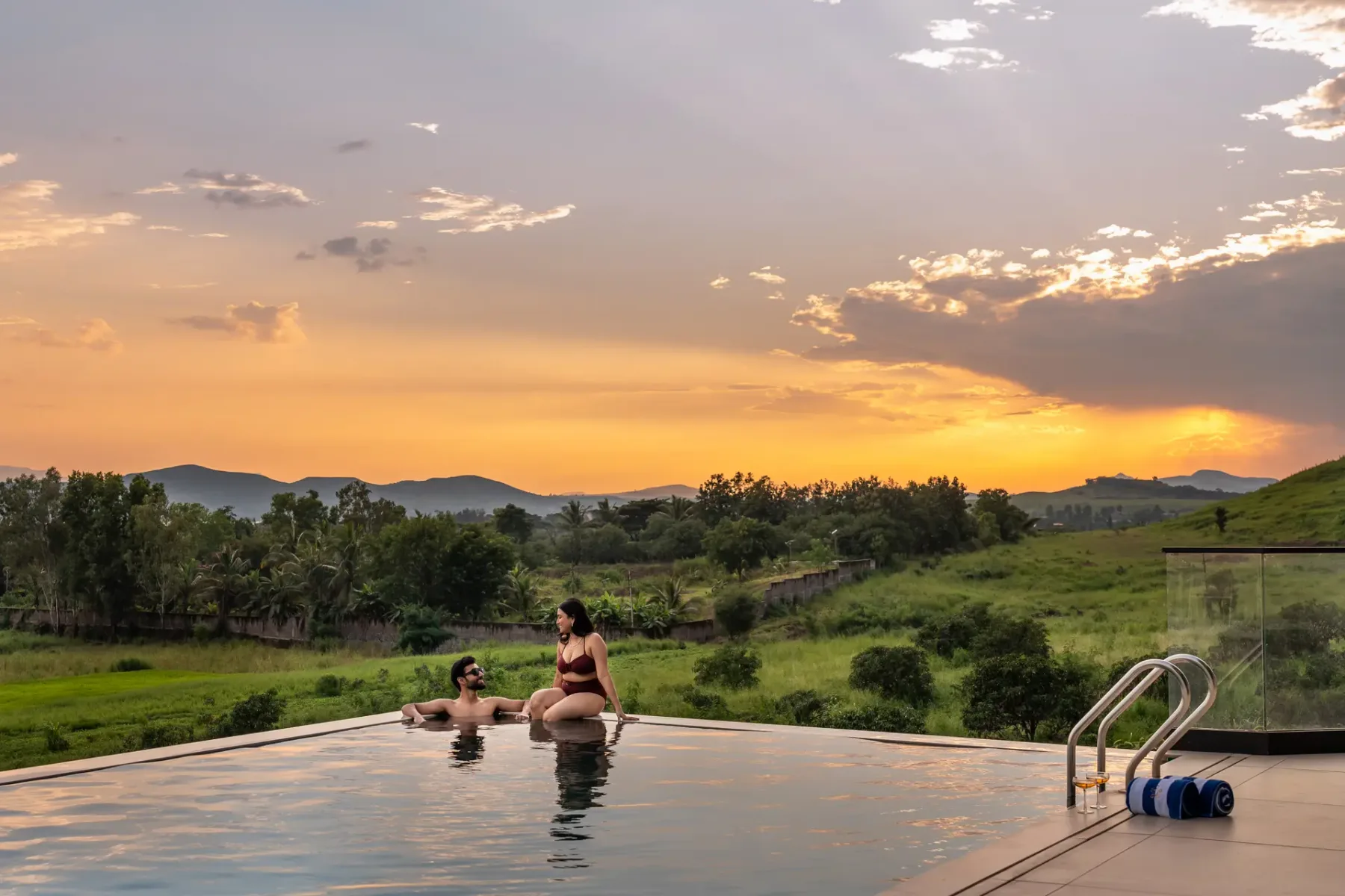 Una piscina al aire libre mira hacia colinas verdes bajo un cielo colorido al atardecer.