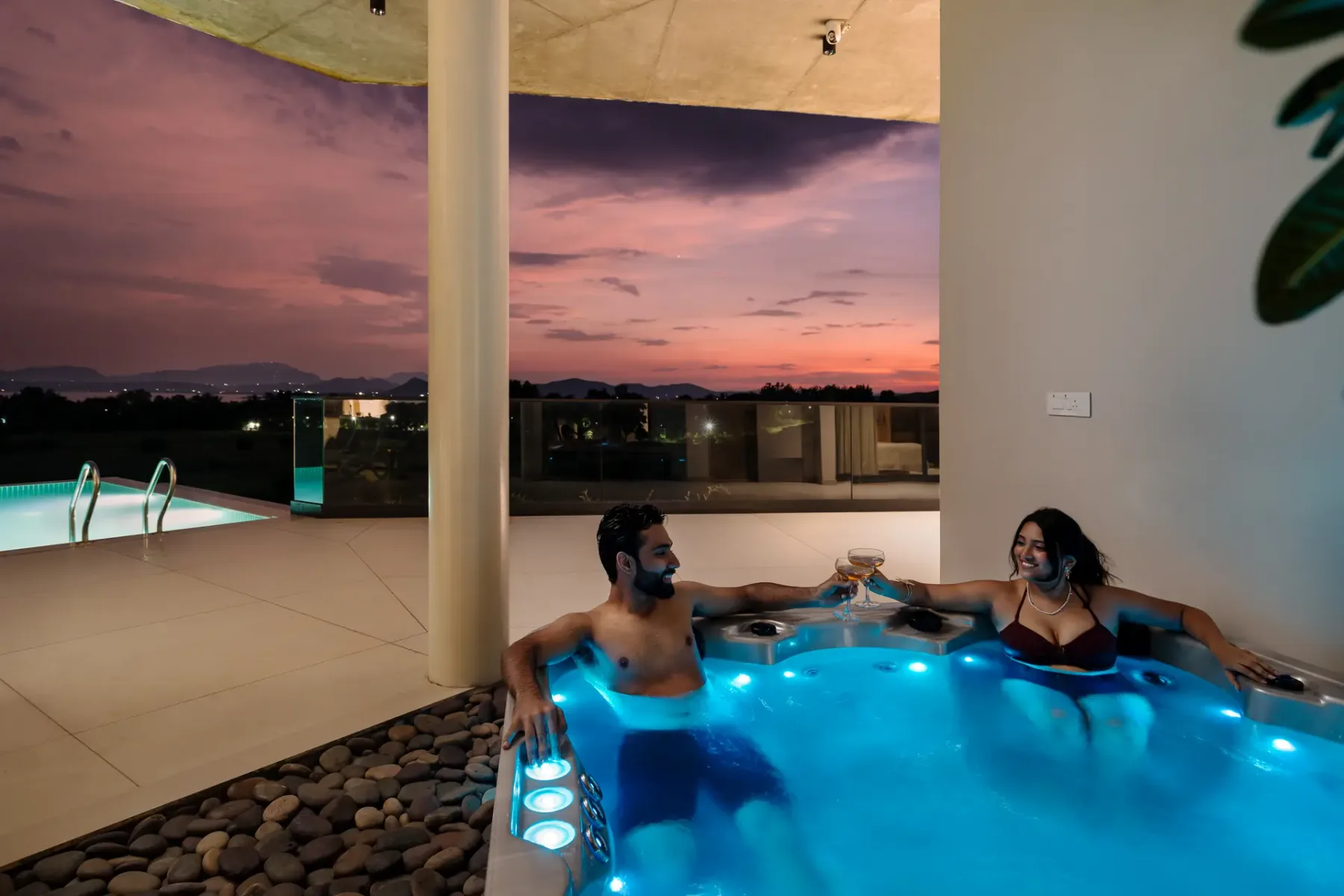 Una pareja se relaja en una bañera de hidromasaje iluminada junto a una piscina al atardecer.