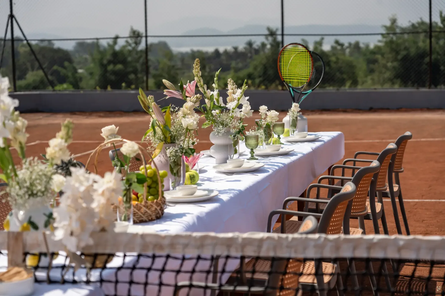 Una mesa al aire libre está preparada junto a una pista de tenis, con arreglos florales.