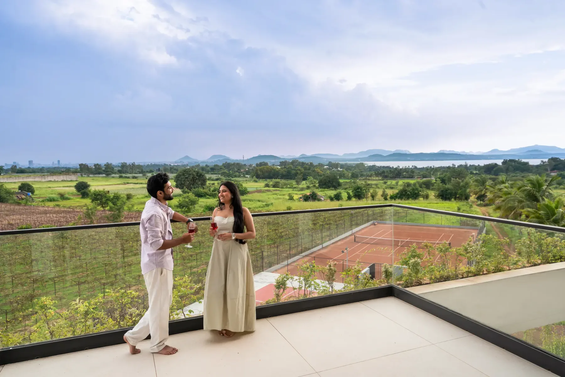 Una pareja está en una terraza con vistas a campos verdes y una cancha de tenis.