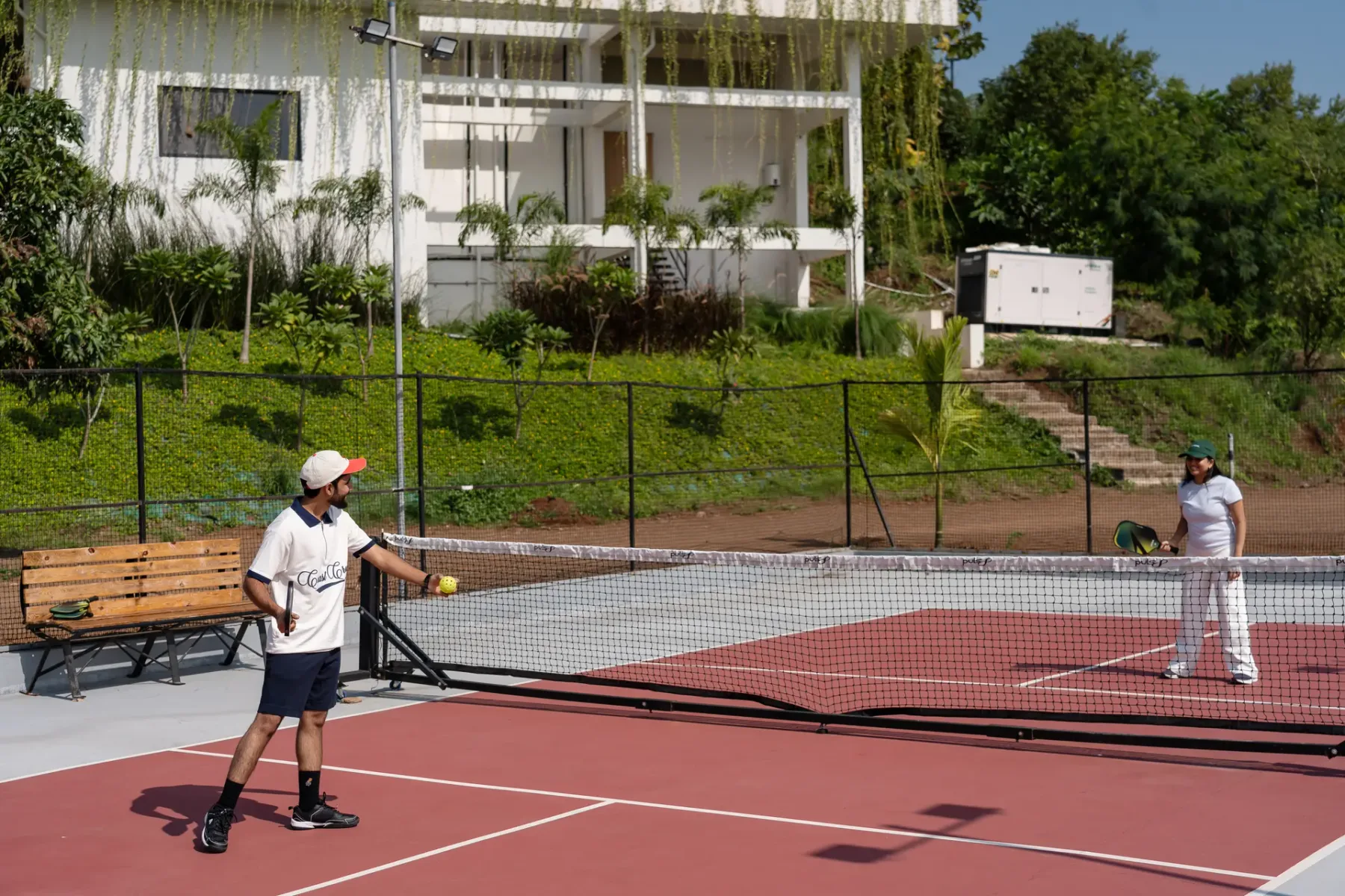 Dos personas juegan al tenis en una cancha al aire libre junto a un edificio blanco.