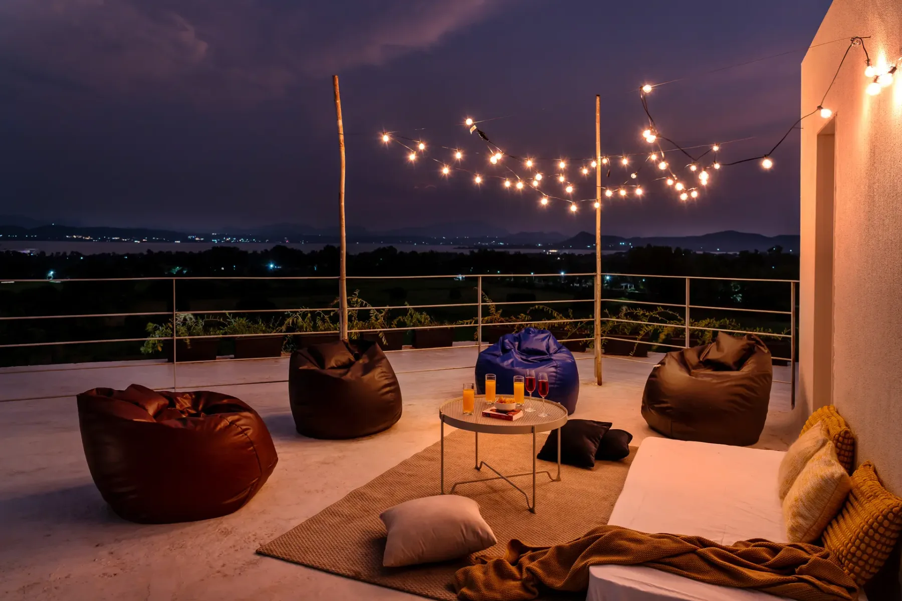 Una terraza al anochecer tiene luces colgantes, asientos informales y vistas de colinas lejanas.