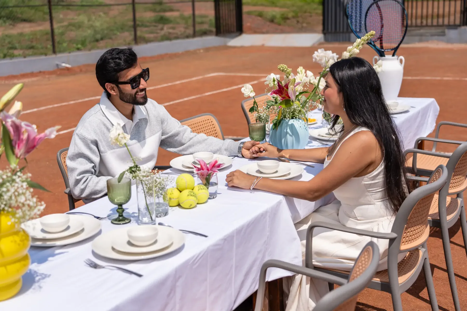 Una pareja comparte una comida en una mesa decorada al aire libre, junto a una cancha de tenis.