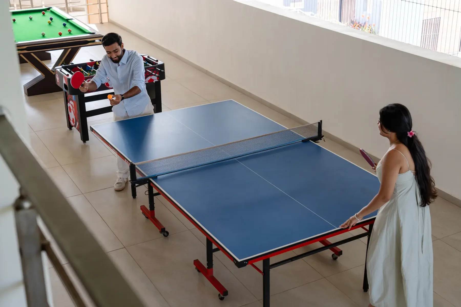 Una sala recreativa con dos personas jugando al tenis de mesa y una mesa de billar cerca.