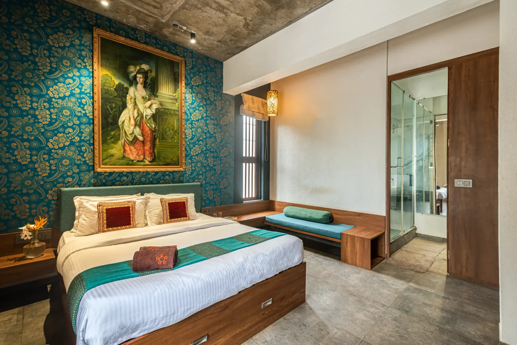 A bedroom with a bed beside a teal upholstered bench and a glass shower area.