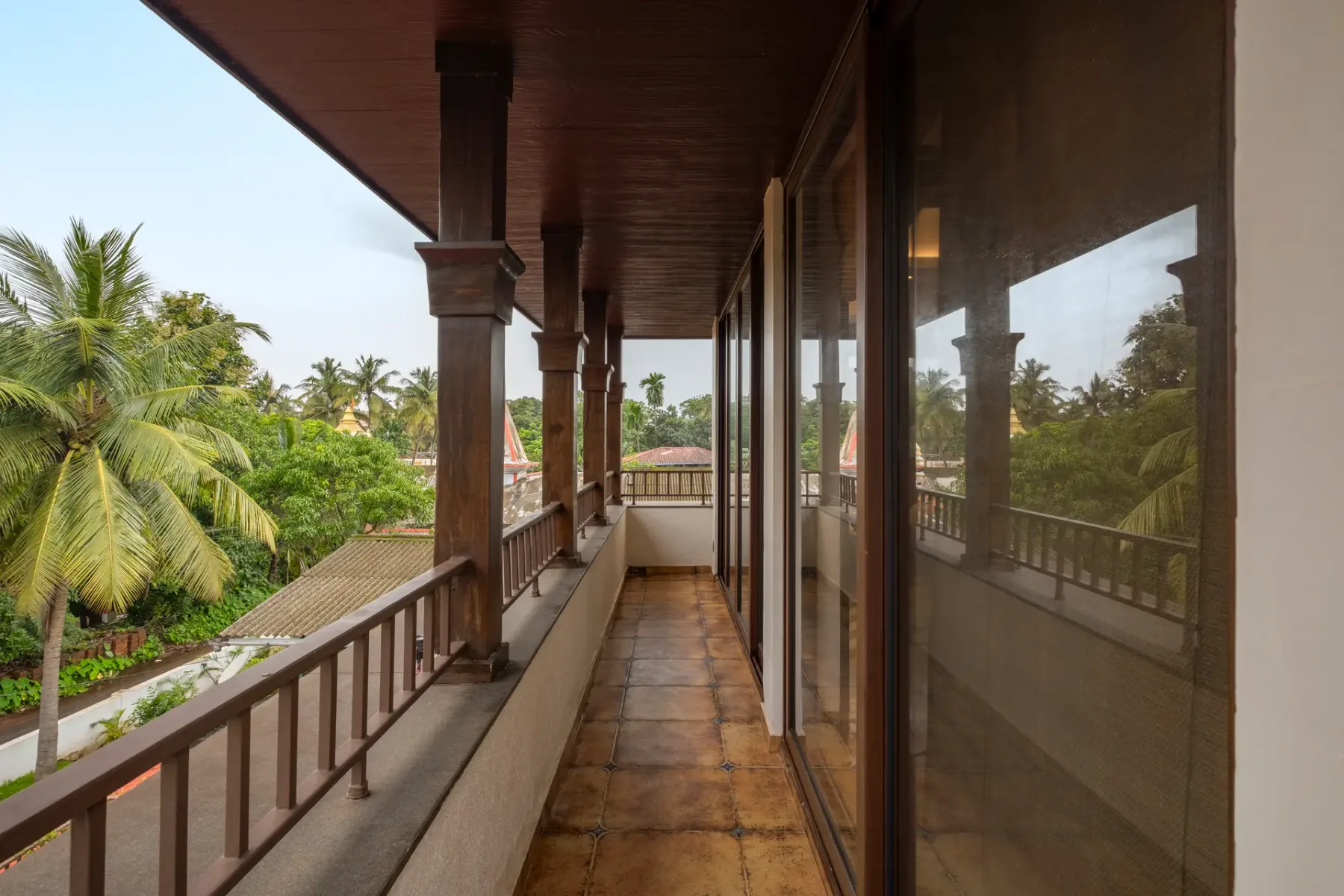 A covered balcony corridor has wooden railings, tiled flooring, and glass doors overlooking palm trees.