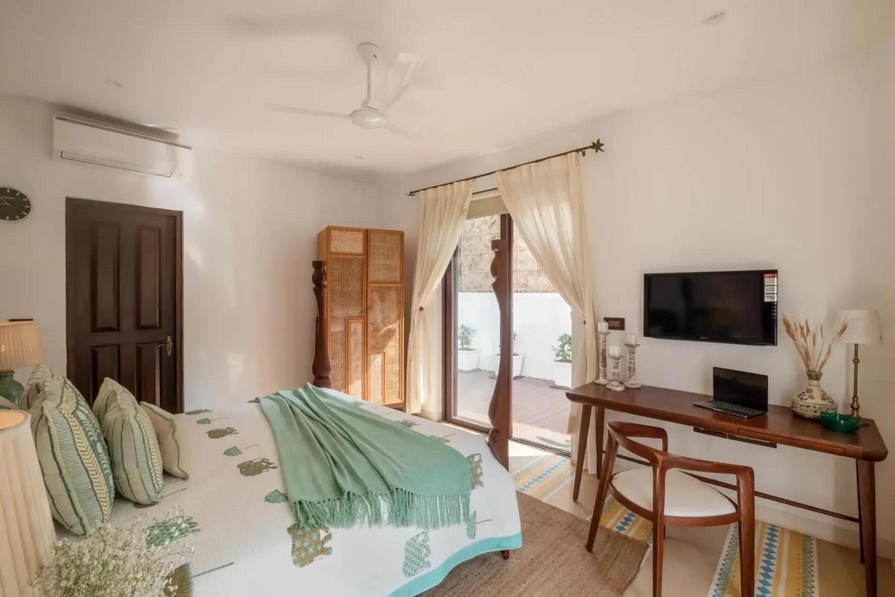 A bedroom with a patterned bed, a writing desk, and glass doors opening onto a small balcony.