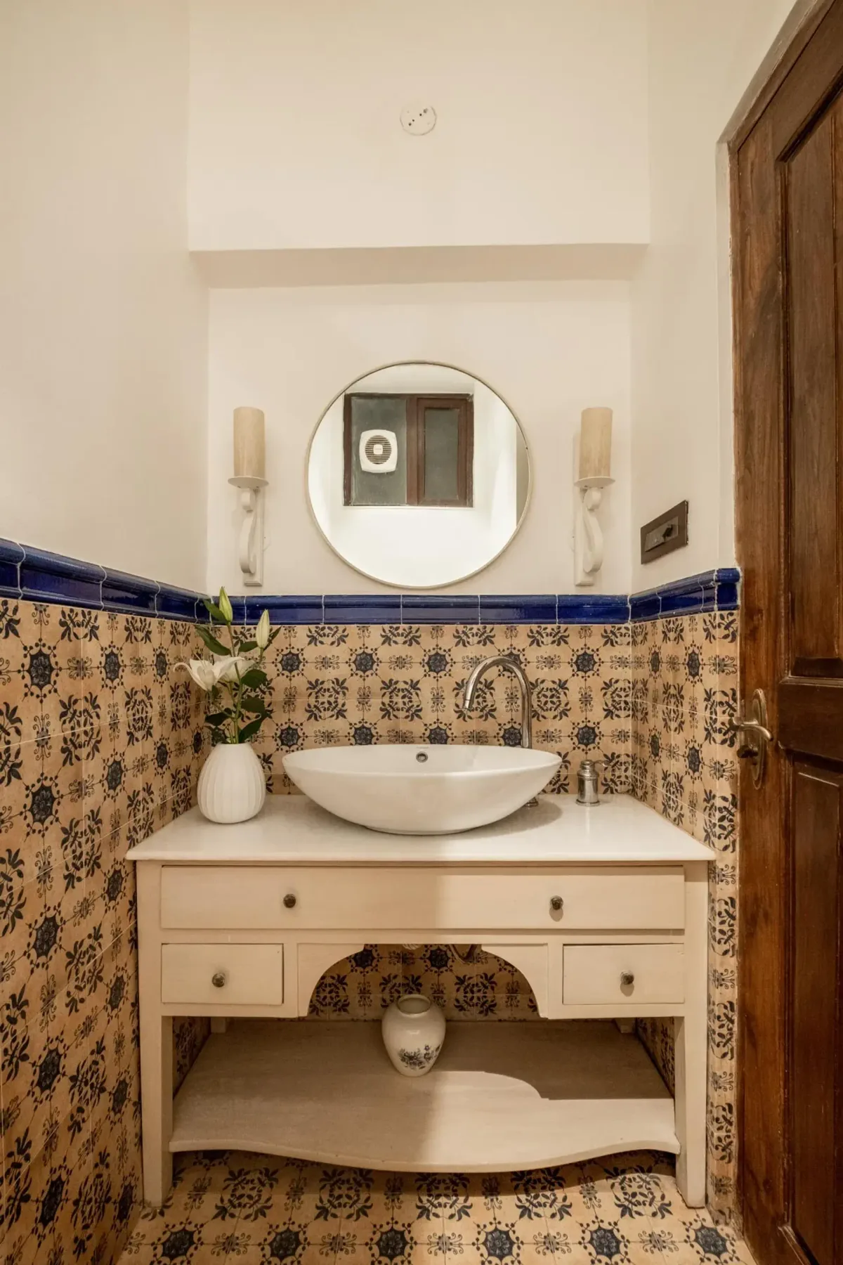 A tiled bathroom vanity has a white vessel sink, round mirror, wall sconces, and a wooden cabinet.