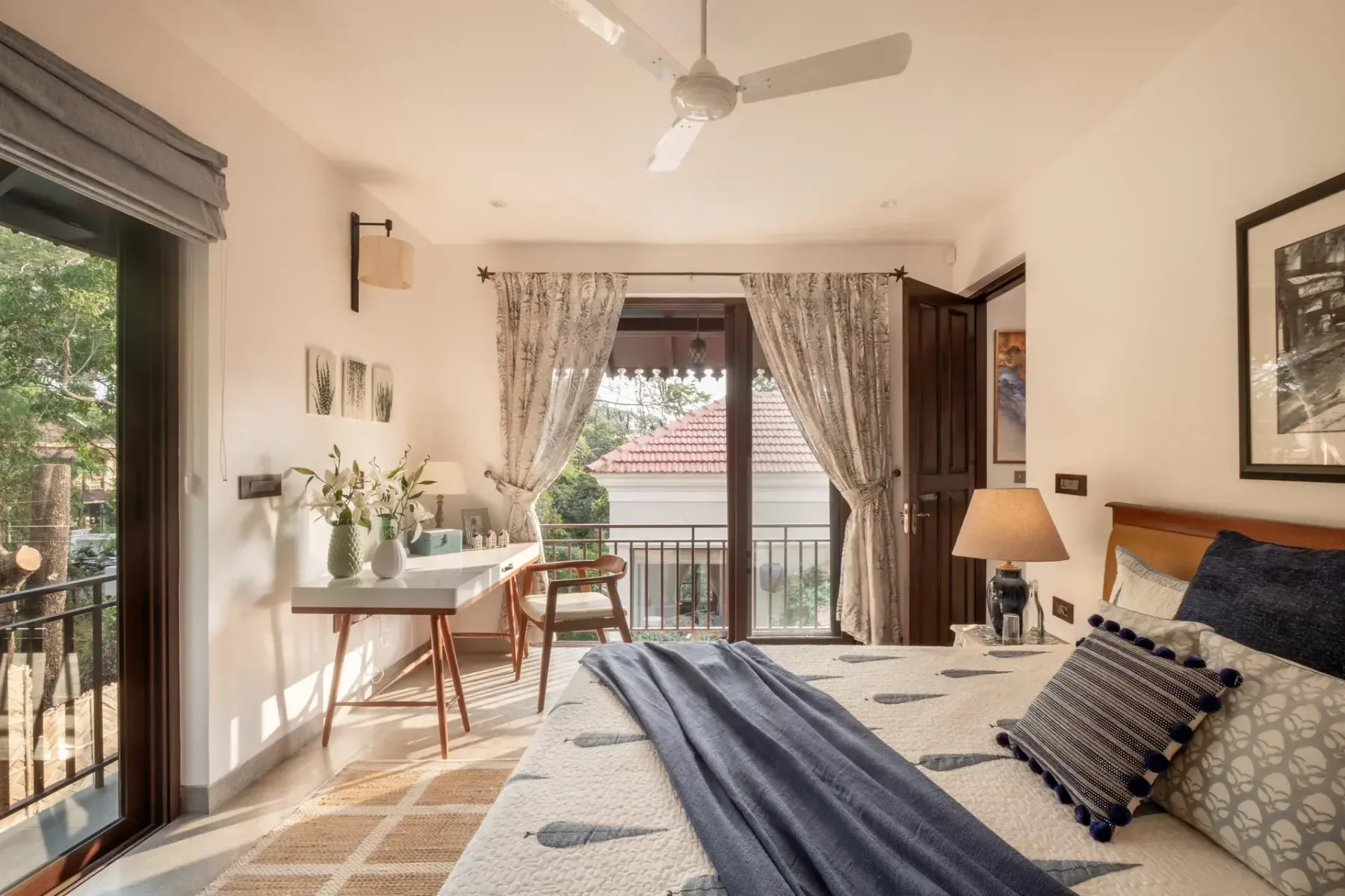 A bright bedroom has a large bed, a writing desk, and open doors leading to a small balcony.