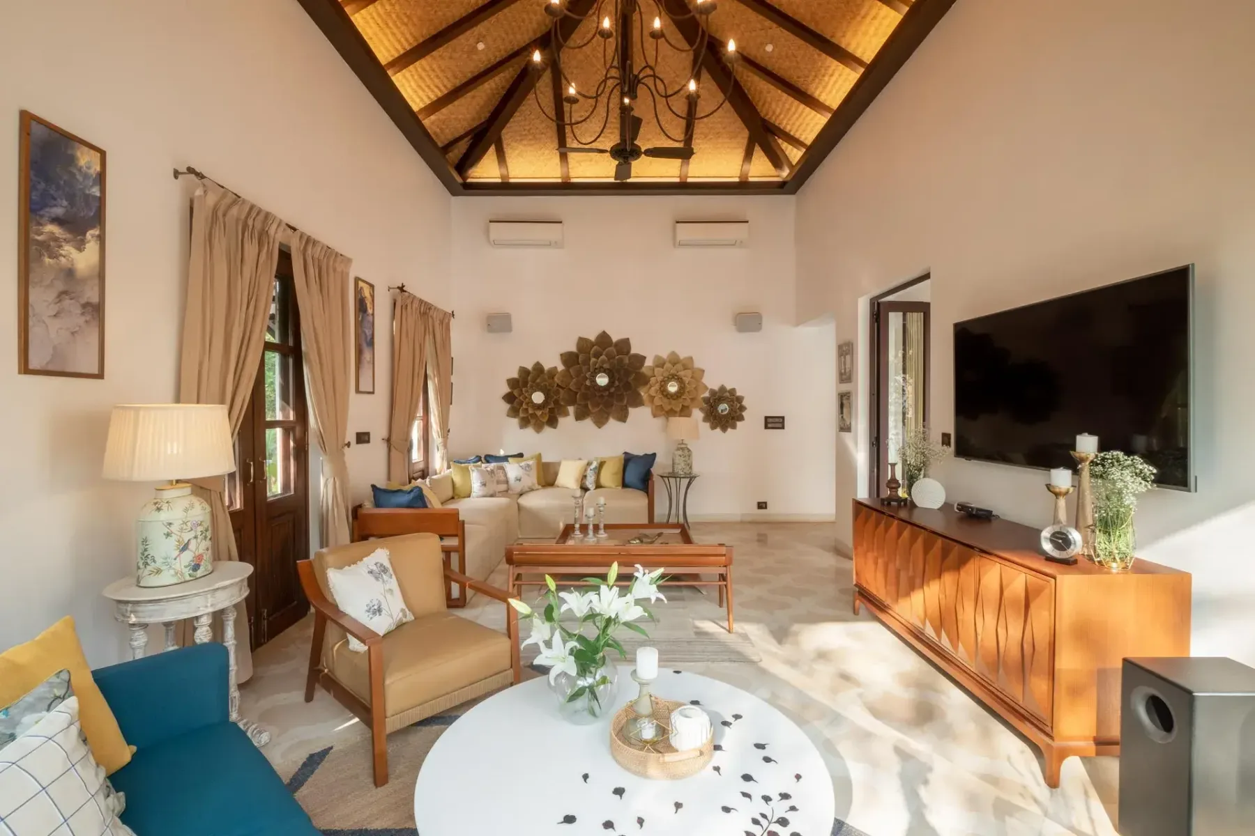 A bright living room has a vaulted timber ceiling, curtained windows, sofas, and a large television.