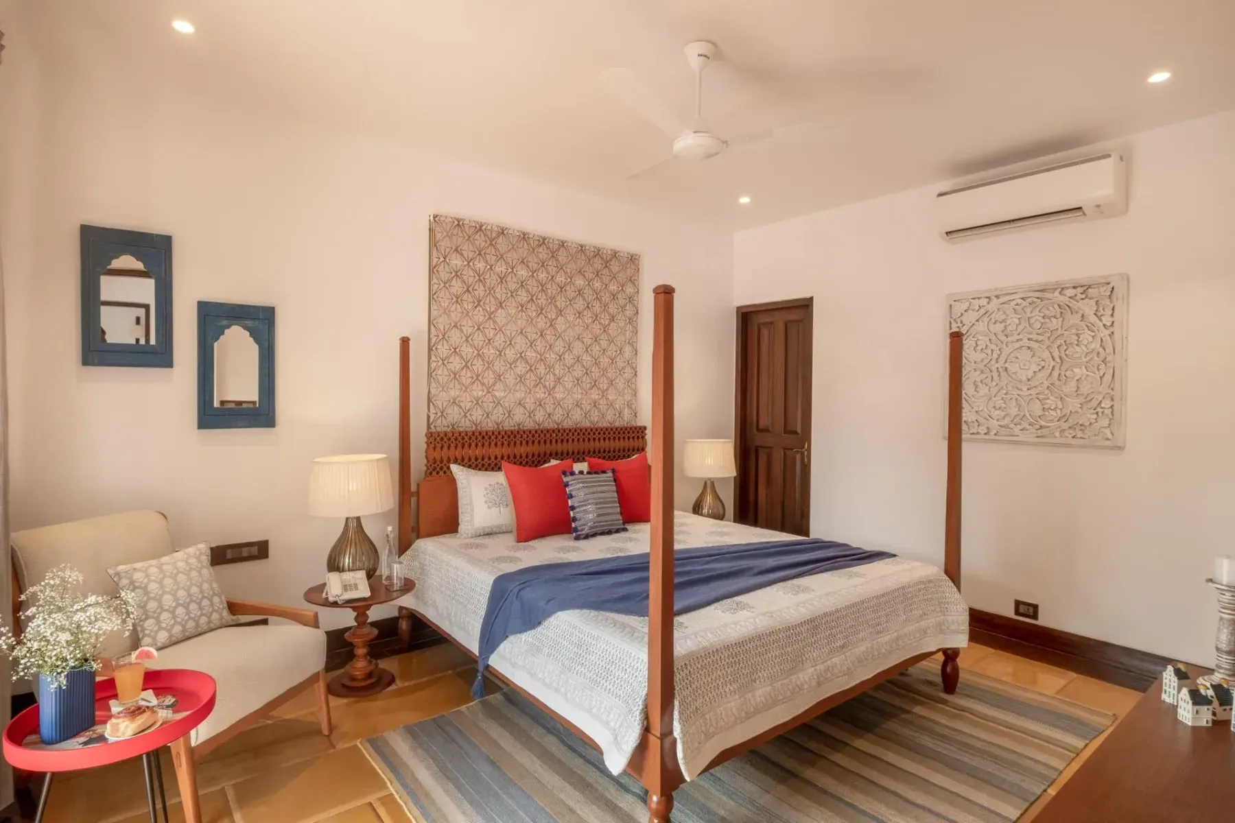 A four-poster bed with blue and red pillows anchors a bright bedroom with patterned wall art and wooden floors.
