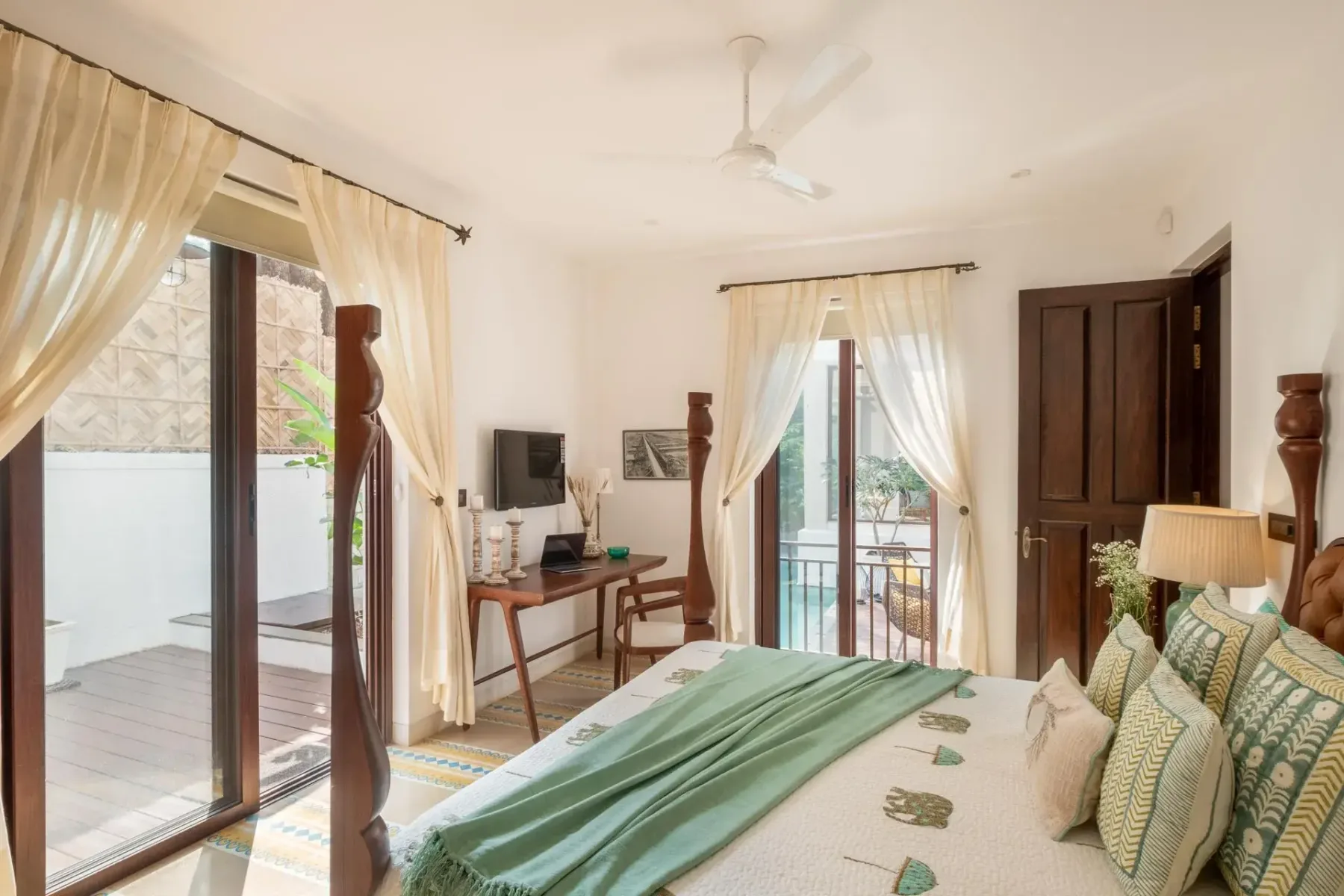 A bright bedroom with a green-covered bed, curtained glass doors, a writing desk, and a ceiling fan.