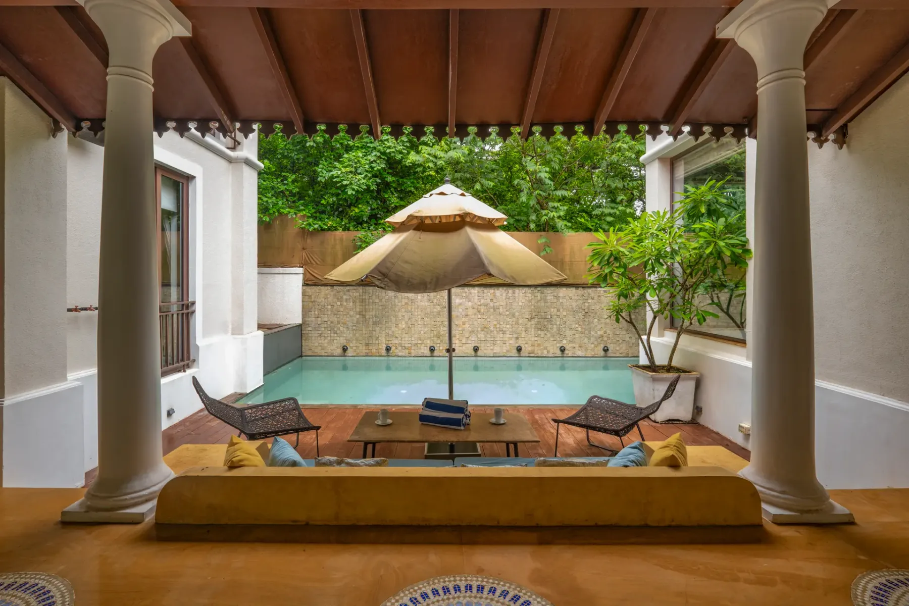 A small courtyard pool sits beneath a wooden pergola, with a central umbrella and seating.