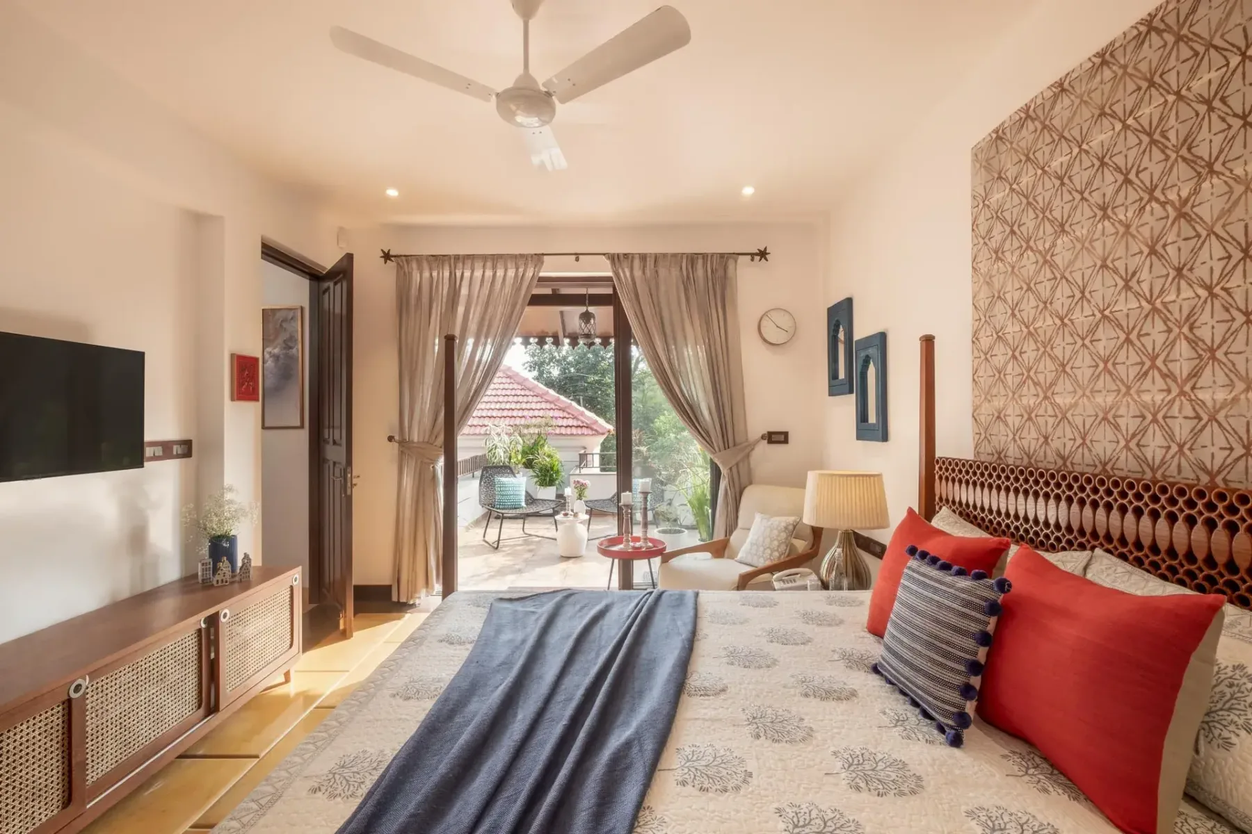 A bright bedroom opens onto a balcony, with a large bed, patterned headboard, and ceiling fan.
