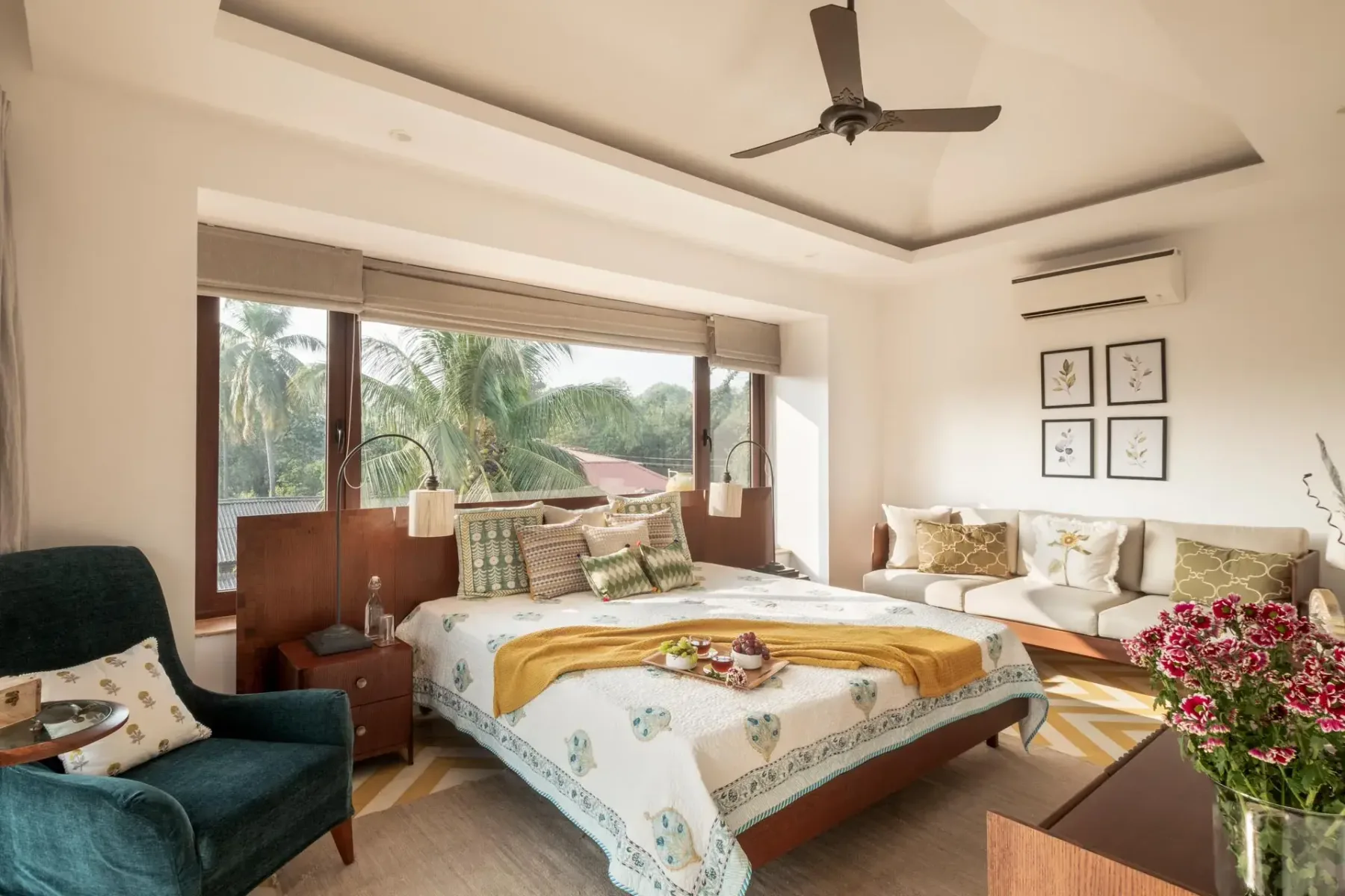 A bright bedroom features a large bed, a teal armchair, and a cream sofa beside broad windows.