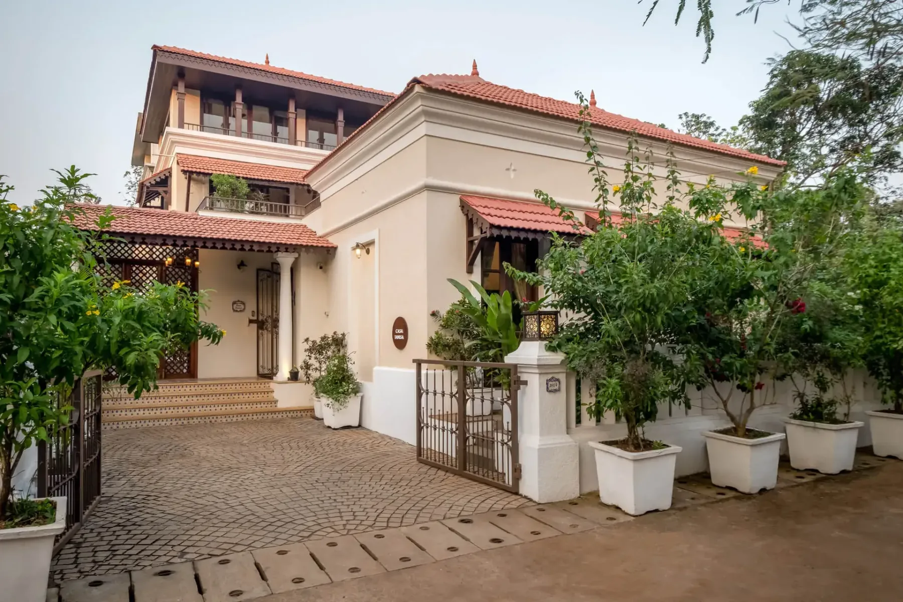 A cream-colored house with red tile roofs, a paved courtyard, and potted trees by the entrance.