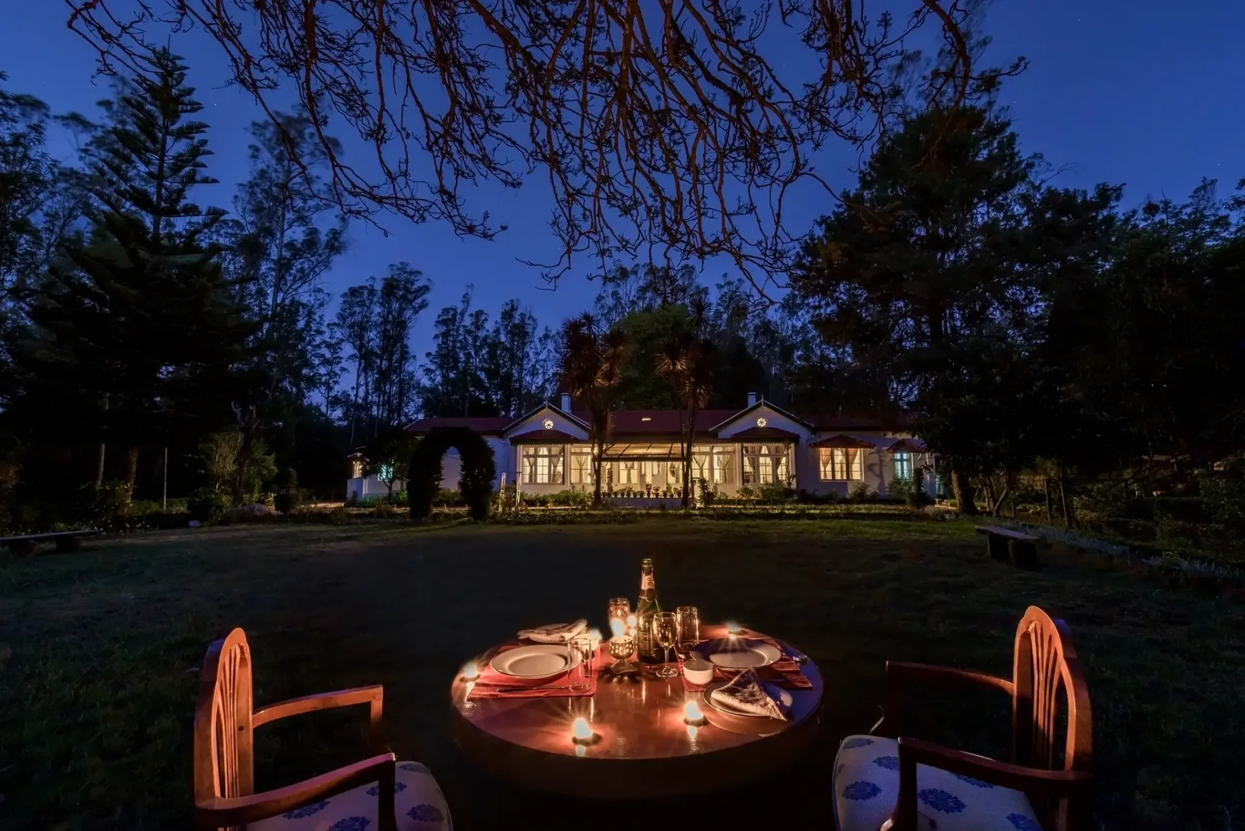 Una mesa al aire libre con velas está en un jardín frente a una casa iluminada al anochecer.