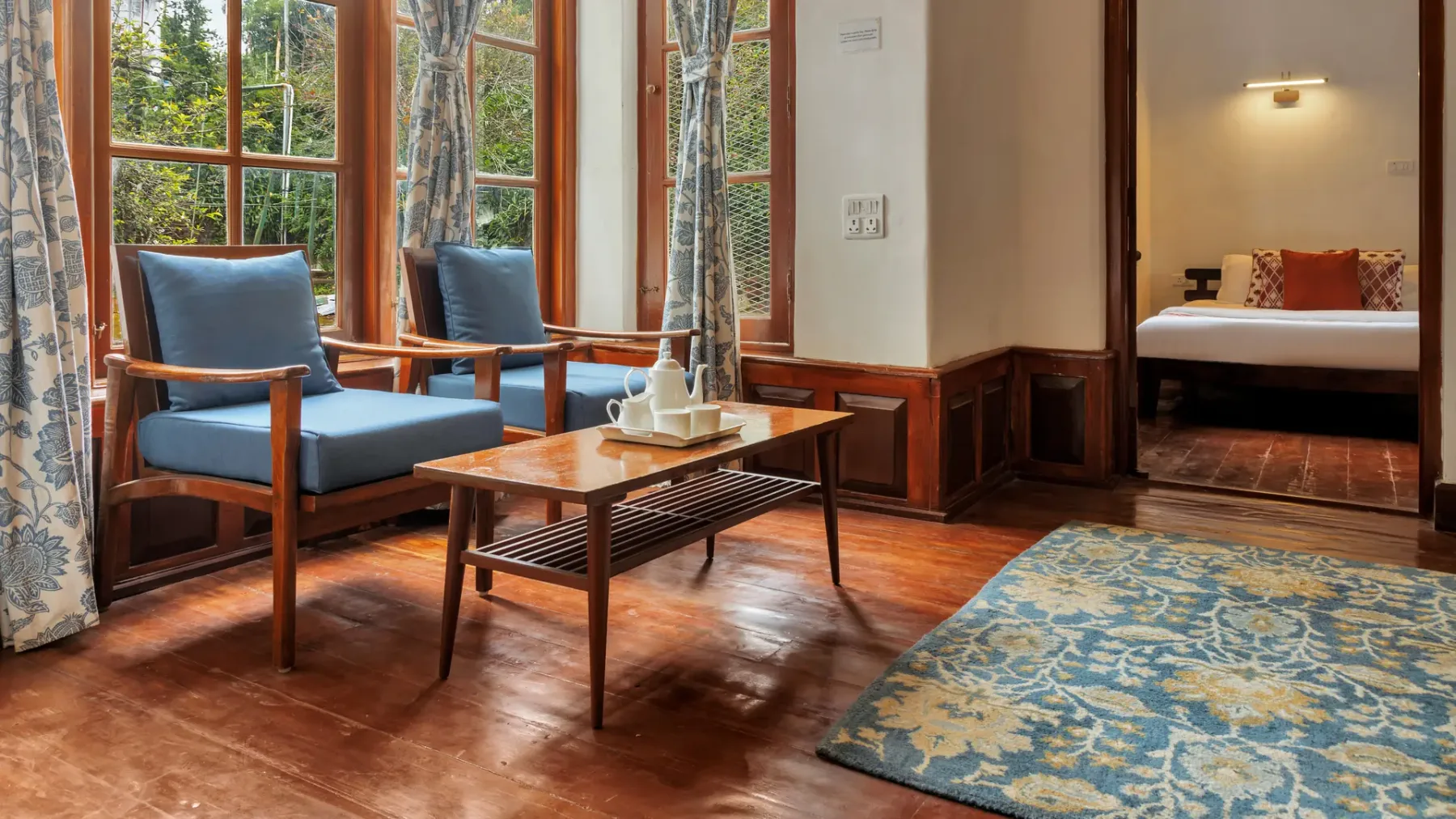 Zona de estar con asientos azules, una mesa de madera y una alfombra estampada junto a grandes ventanas.