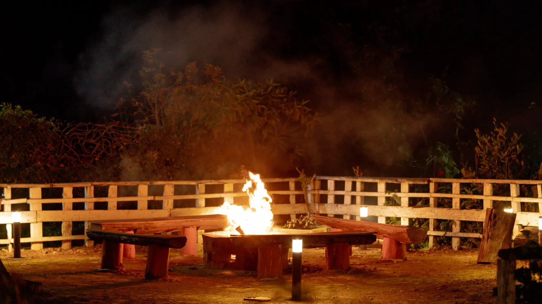 Una fogata al aire libre arde junto a una barandilla de madera por la noche.
