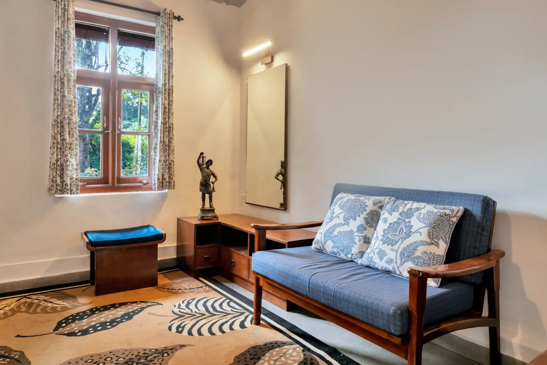 Una zona de estar con asiento azul, una ventana y una alfombra estampada.