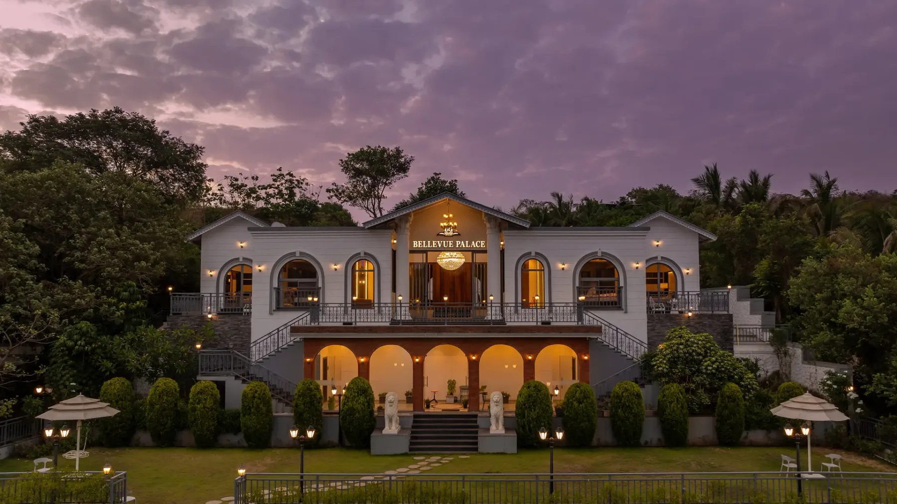 A large illuminated building stands among trees beneath a cloudy evening sky.