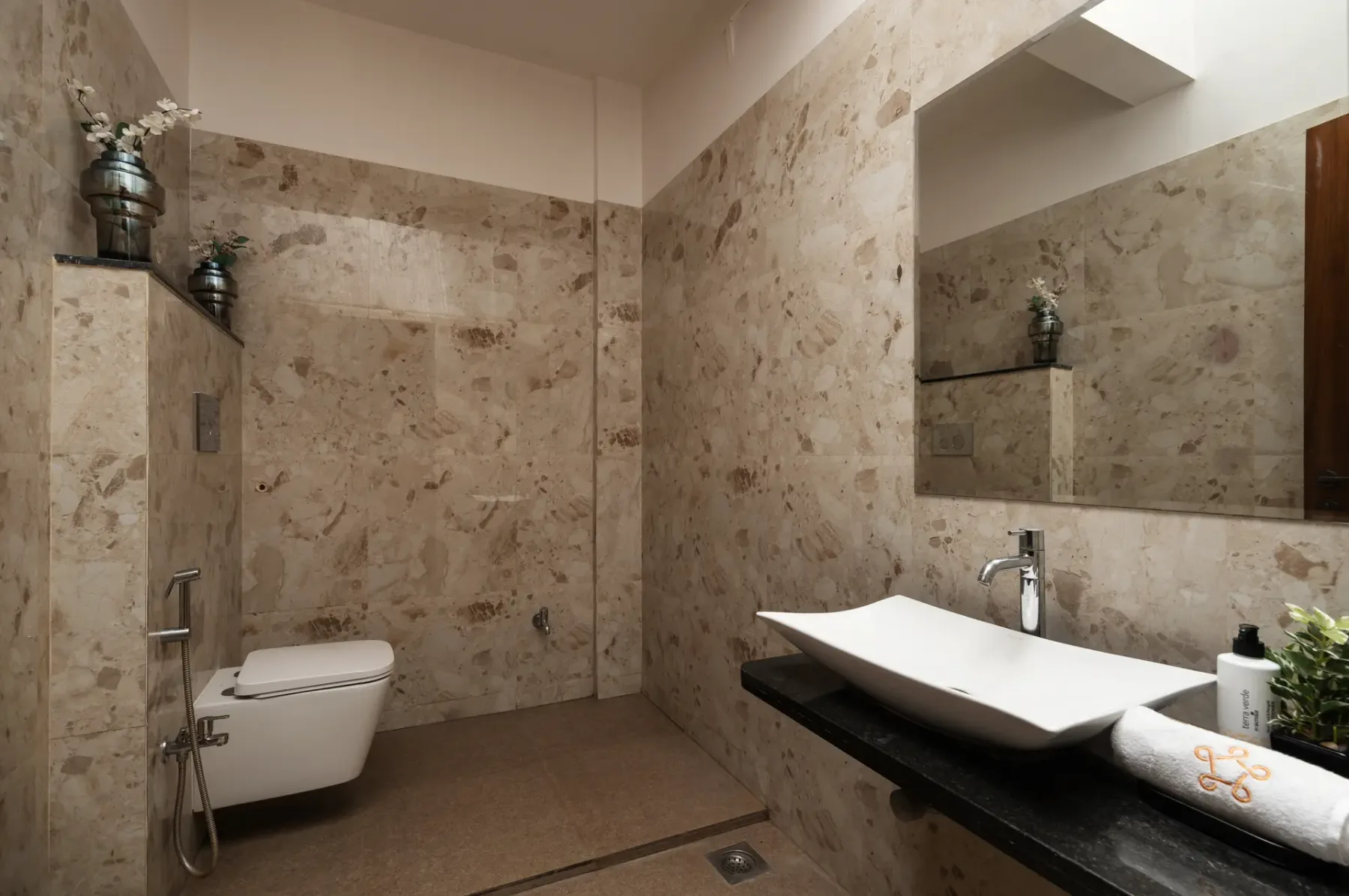 A tiled bathroom with a wall-mounted toilet and a wide sink beneath a mirror.
