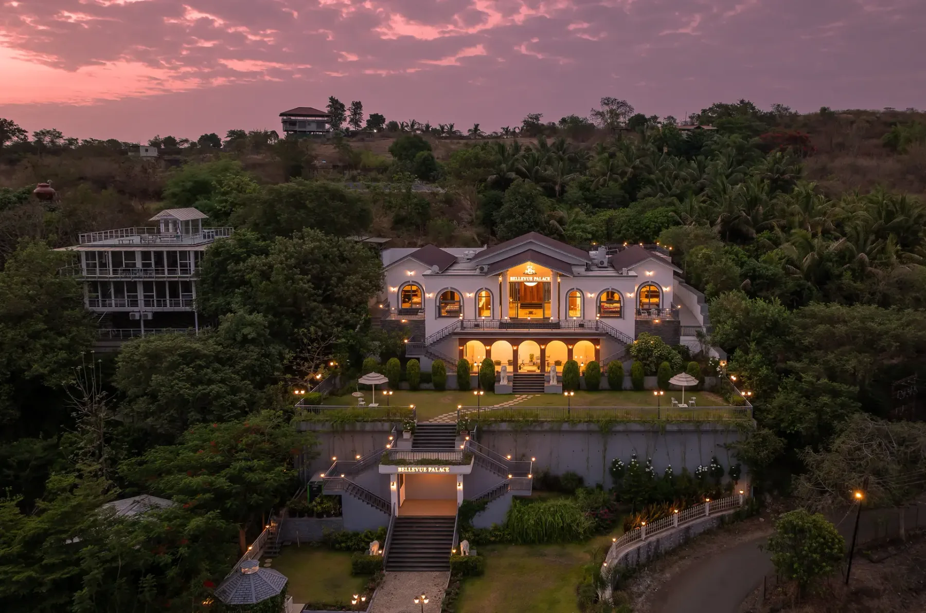 An illuminated estate building sits amid landscaped grounds beneath a pink evening sky.