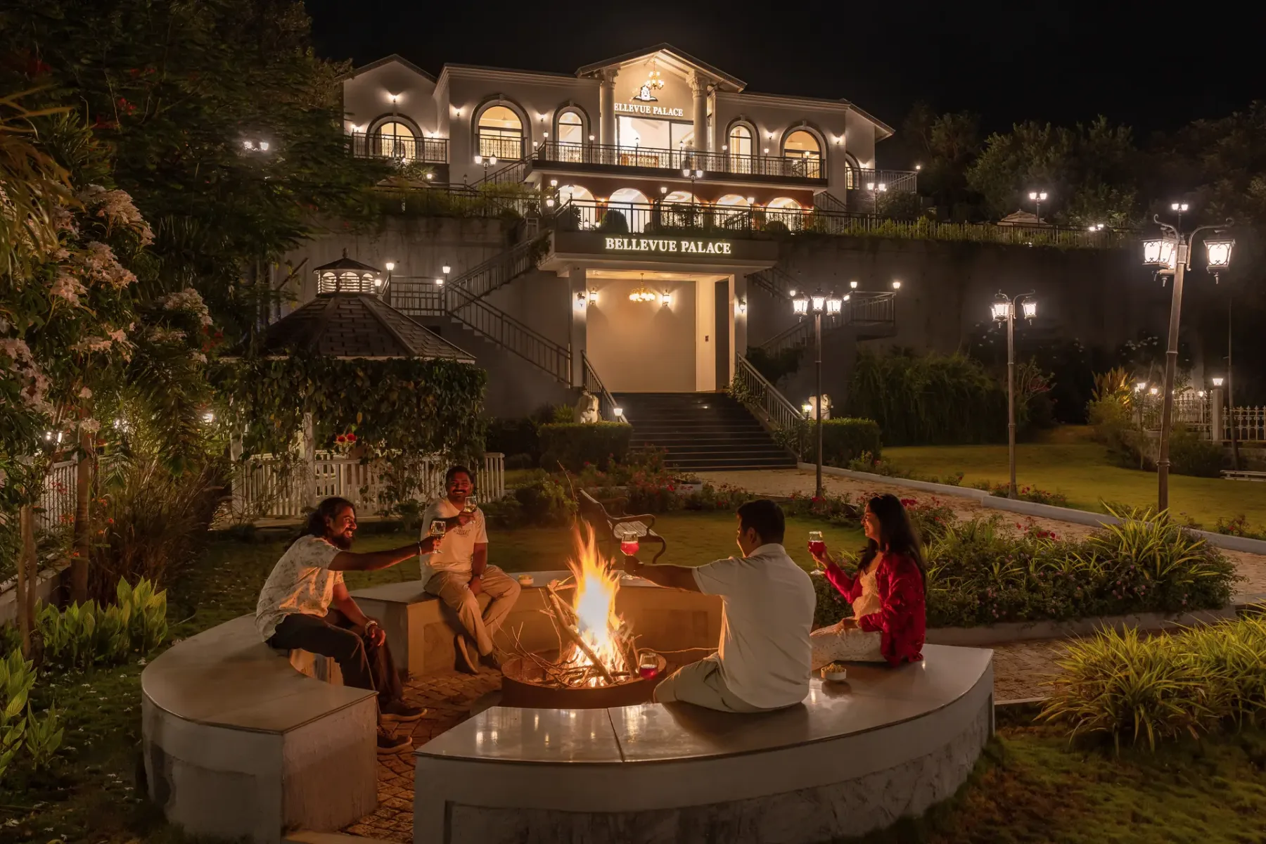 A landscaped outdoor fire pit seating area sits below a brightly lit building at night.
