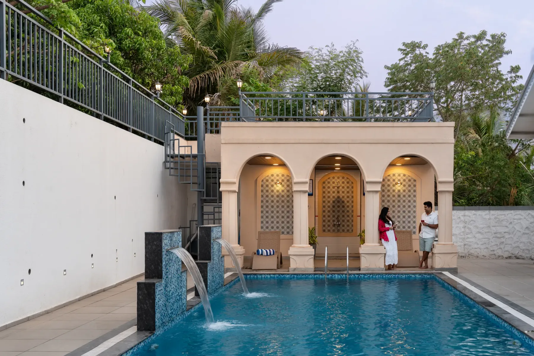 An outdoor swimming pool sits beside a pale building with three arched openings.