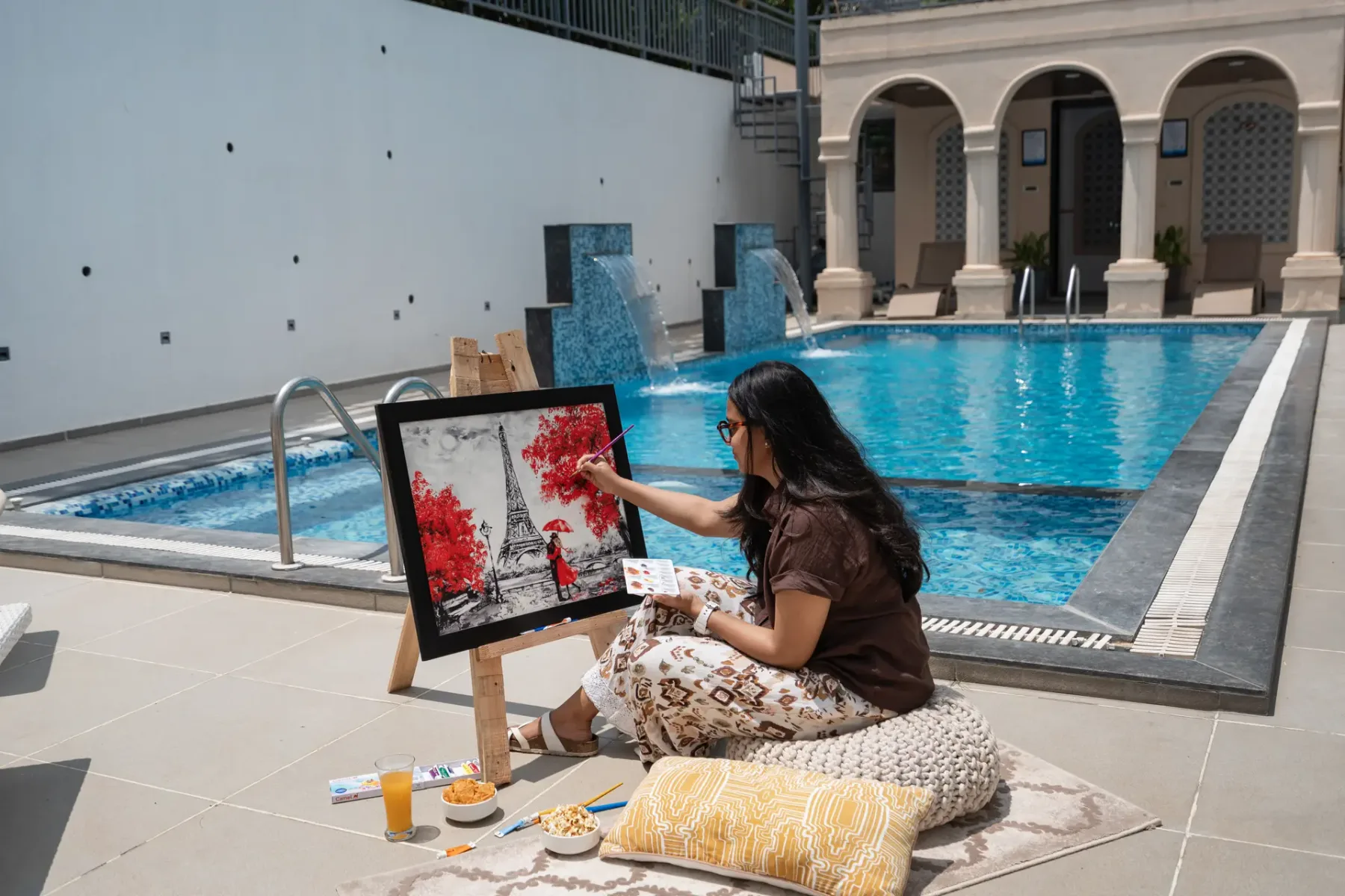 A woman paints beside a blue swimming pool in a tiled courtyard.