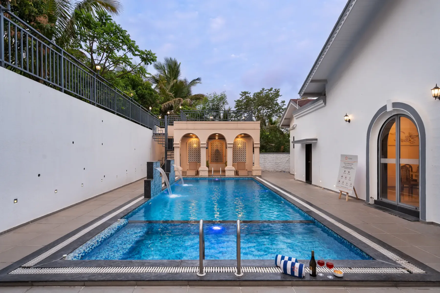 An outdoor swimming pool sits between white buildings, with palm trees in the background.
