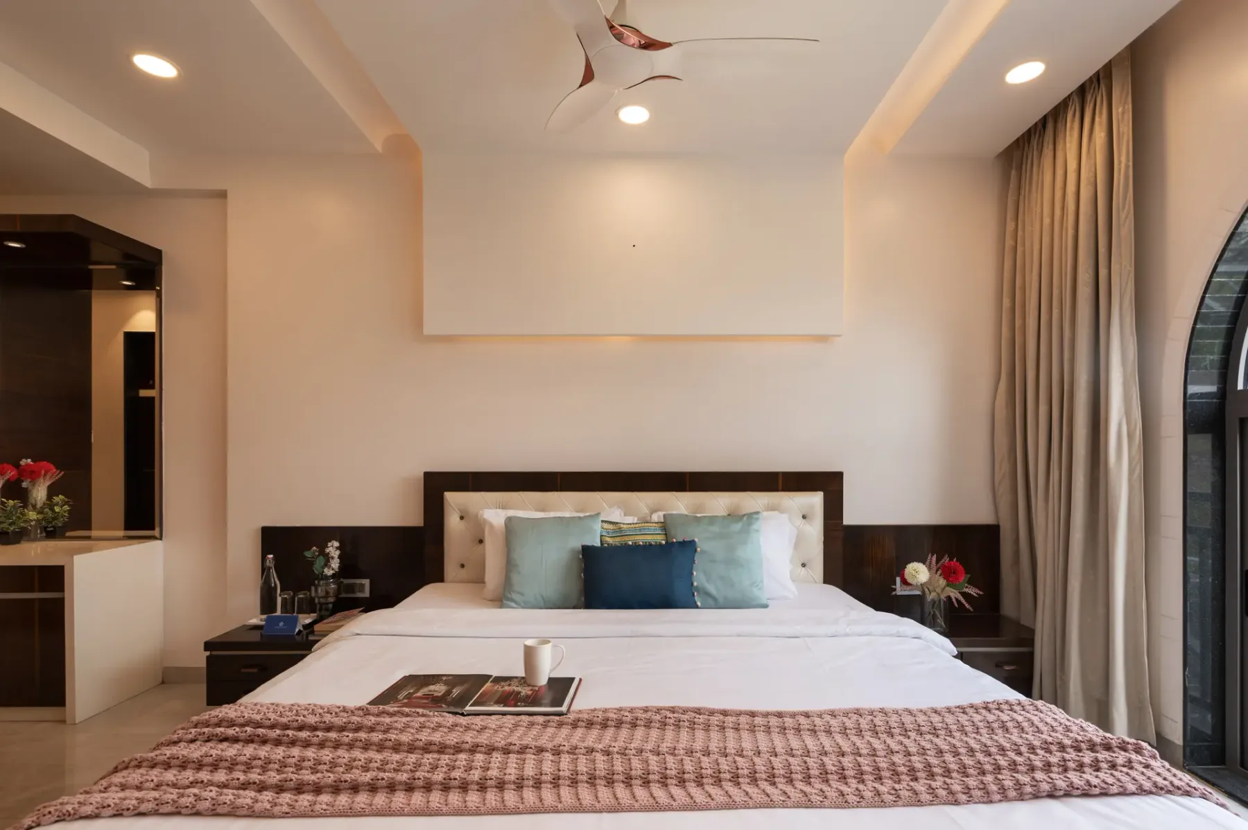 A bedroom with a neatly made bed, colorful pillows, and curtains beside an arched window.