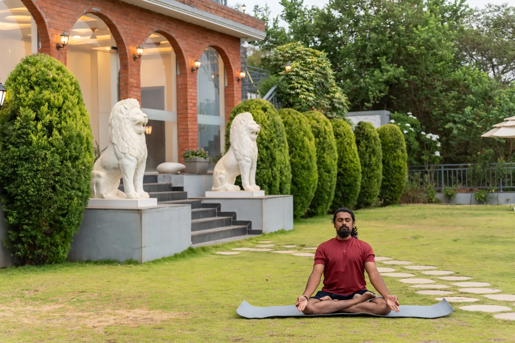 A person sits cross-legged on a mat in a landscaped garden beside an arched building.