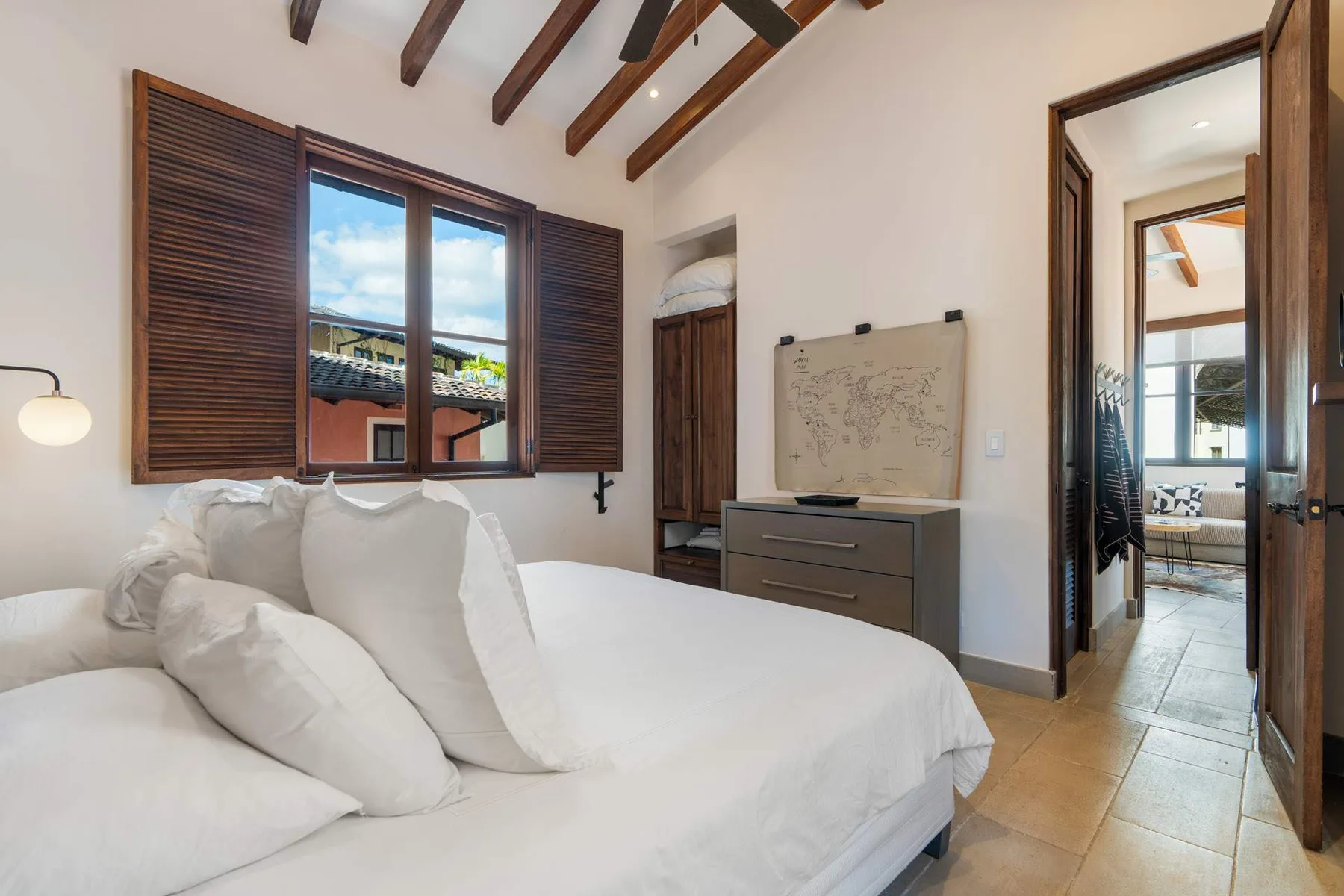 A bright bedroom with a large bed, shuttered window, and exposed ceiling beams.
