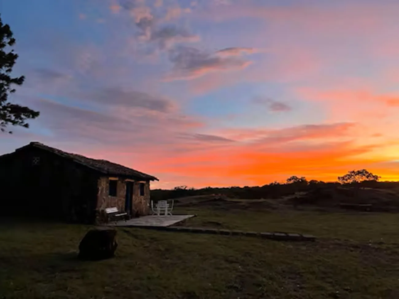 A small cabin sits beside an open grassy landscape beneath a colorful sunset sky.