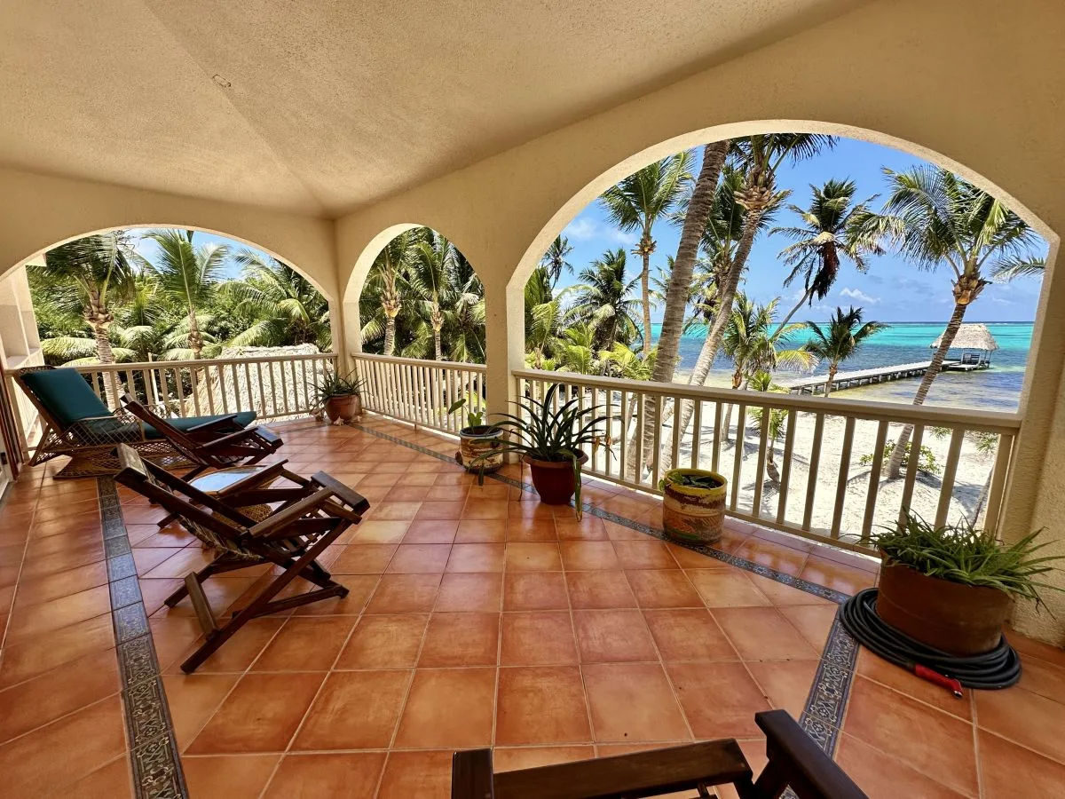 A covered tiled terrace with lounge seating overlooks palm trees and the ocean.