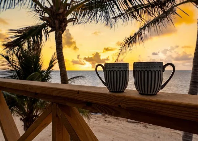 Two mugs sit on a wooden railing overlooking a beach at sunset.