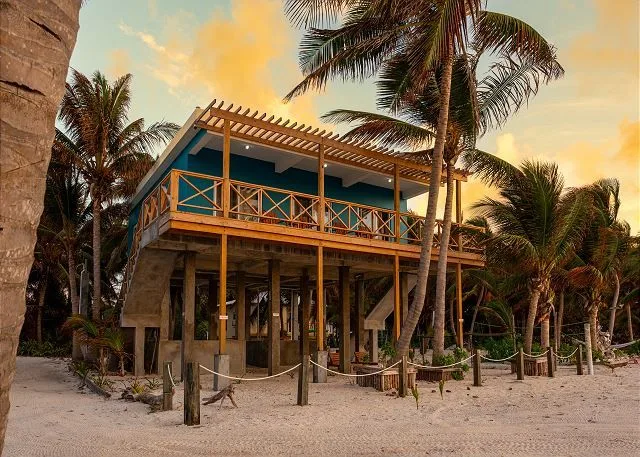 A raised tropical building with a wooden balcony stands among palm trees.
