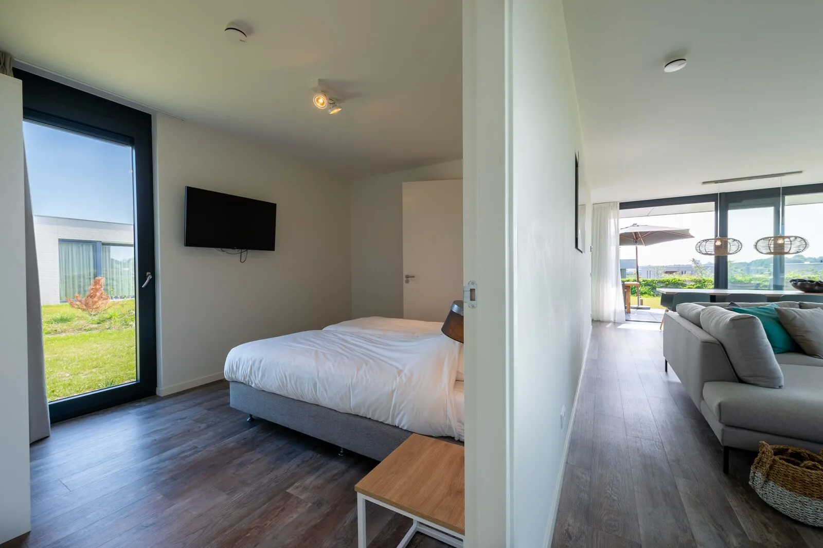 A bedroom with a white bed, wall-mounted television, and glass door opening onto a lawn.