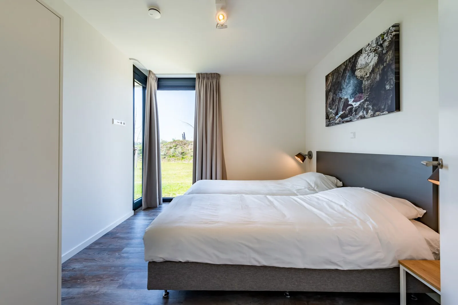 A bedroom with a double bed, white bedding, gray curtains, and a window overlooking a grassy area.