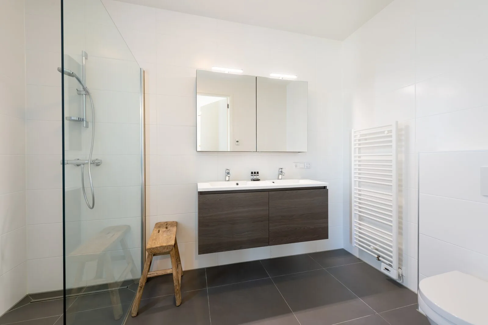 A bright tiled bathroom has a glass shower, a floating dark vanity, and a white towel radiator.