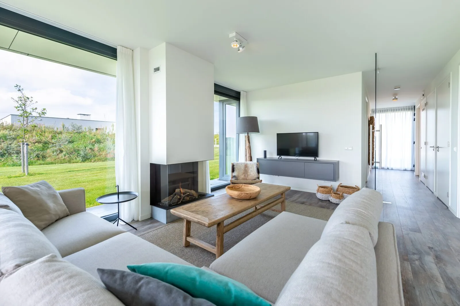 A bright living room with pale sofas, a wooden coffee table, a television, and large windows overlooking a garden.