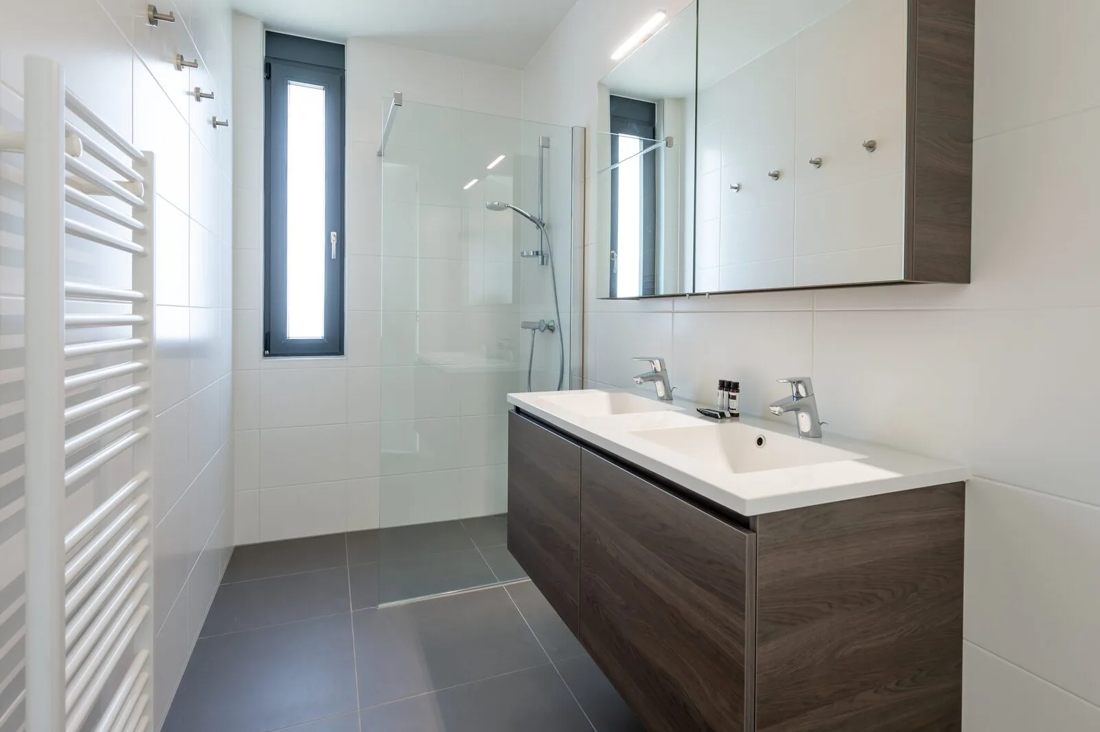 A bright modern bathroom has a glass shower, a double-sink vanity, and a tall narrow window.