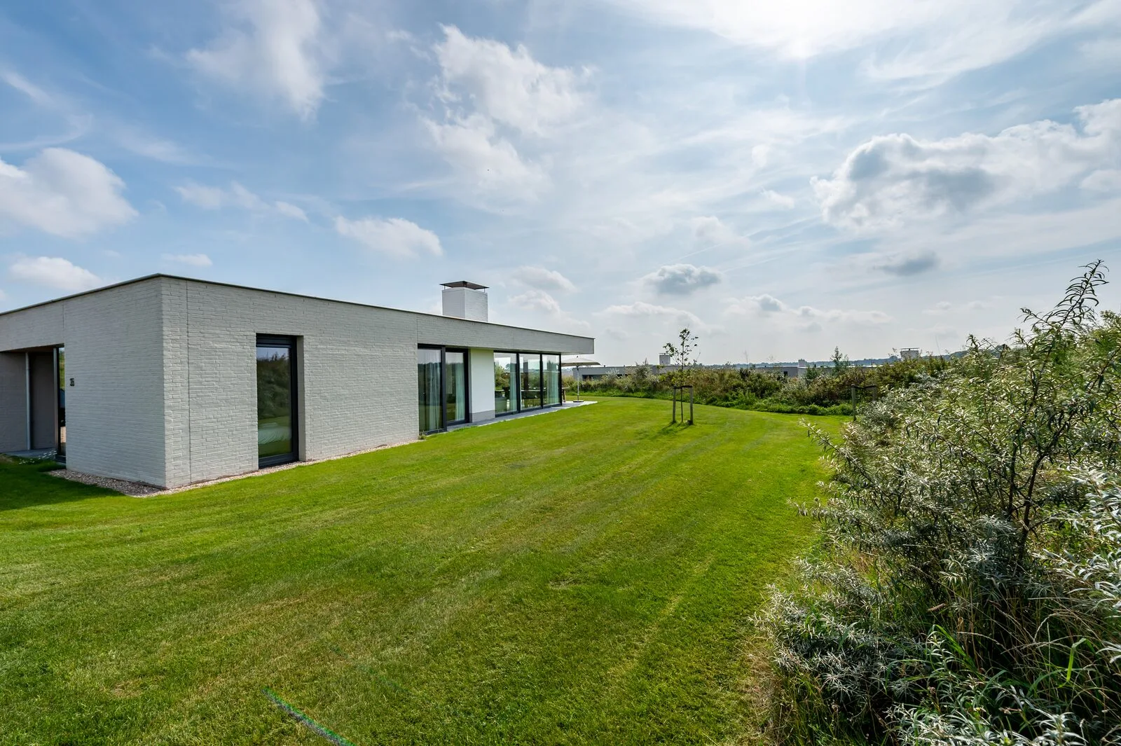 A modern single-story house with broad glass doors stands beside a neatly mown lawn and leafy shrubs.