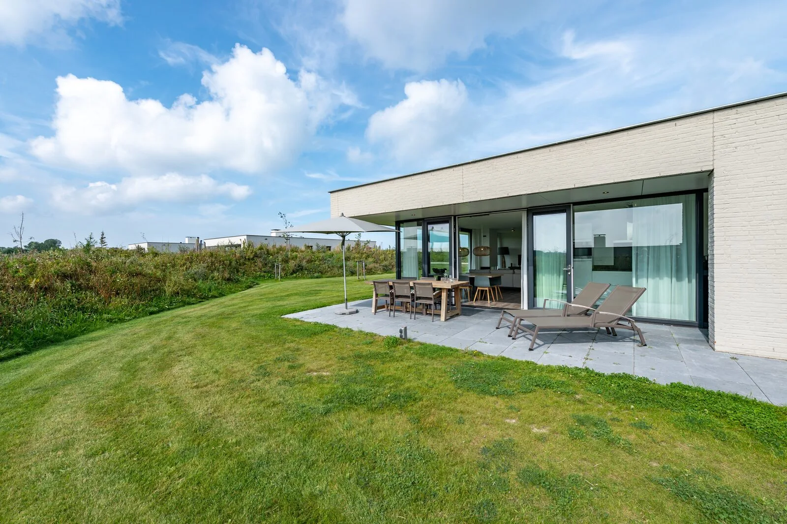 Modern single-story house with broad glass doors, a patio dining set, lounge chairs, and a lawn.
